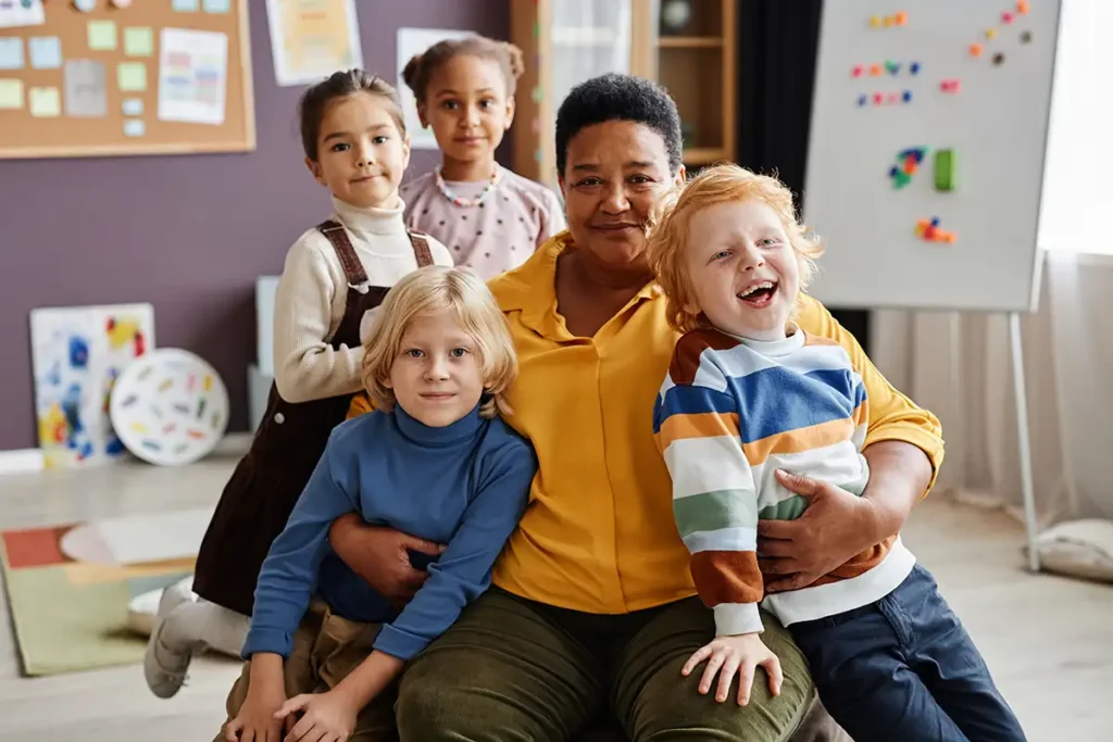 A daycare teacher with a group of smiling children in a classroom.